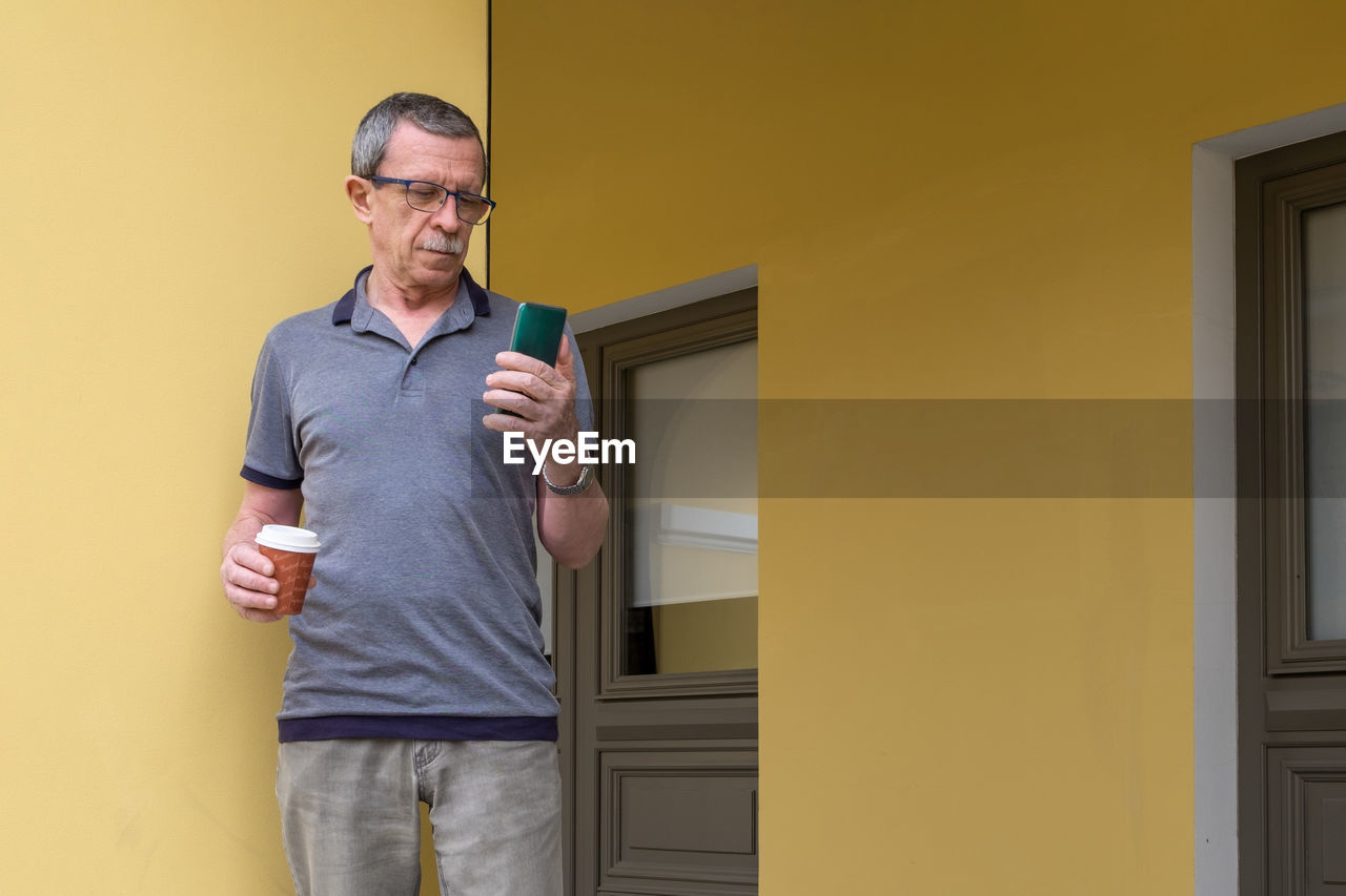 Adult retired man stands against background of yellow wall and holds smartphone and glass of coffee