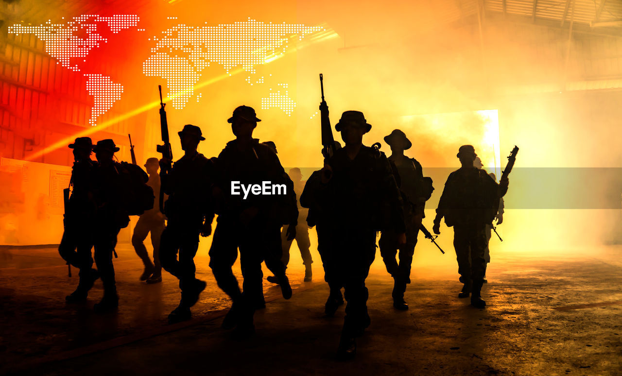 Digital composite image of silhouette soldiers walking with weapon against illuminated world map