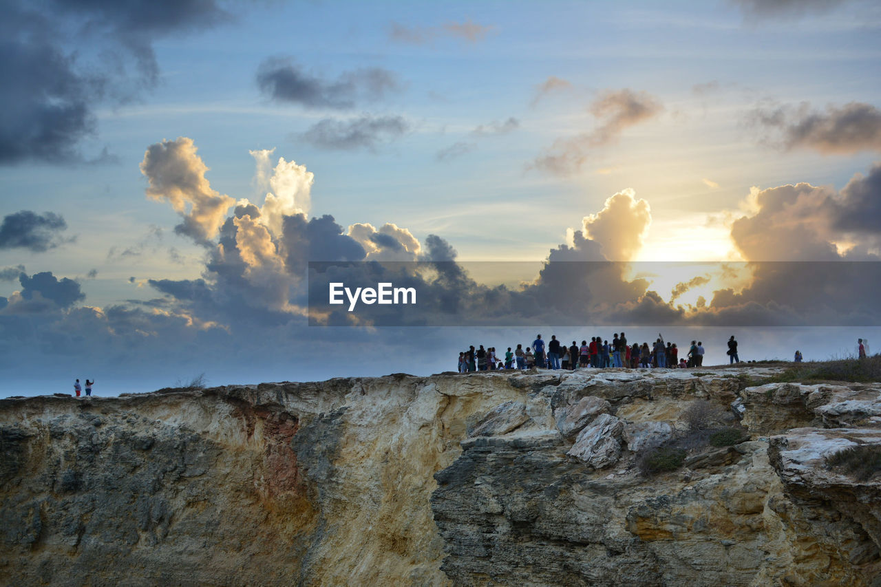 People on rocky mountain against cloudy sky during sunset