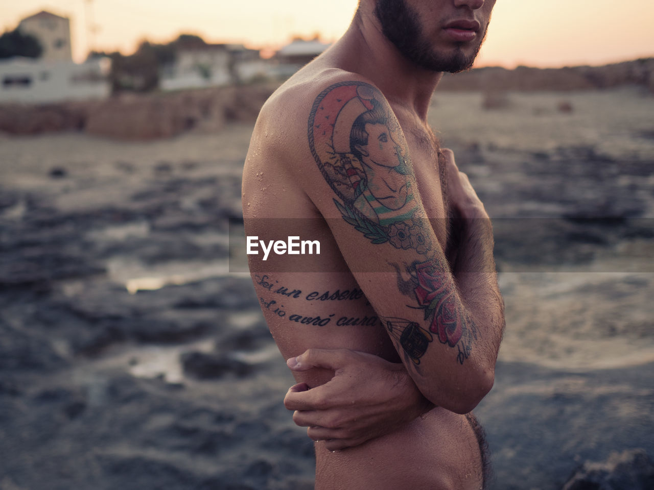 Midsection of shirtless young man with arms crossed standing at beach against sky during sunset