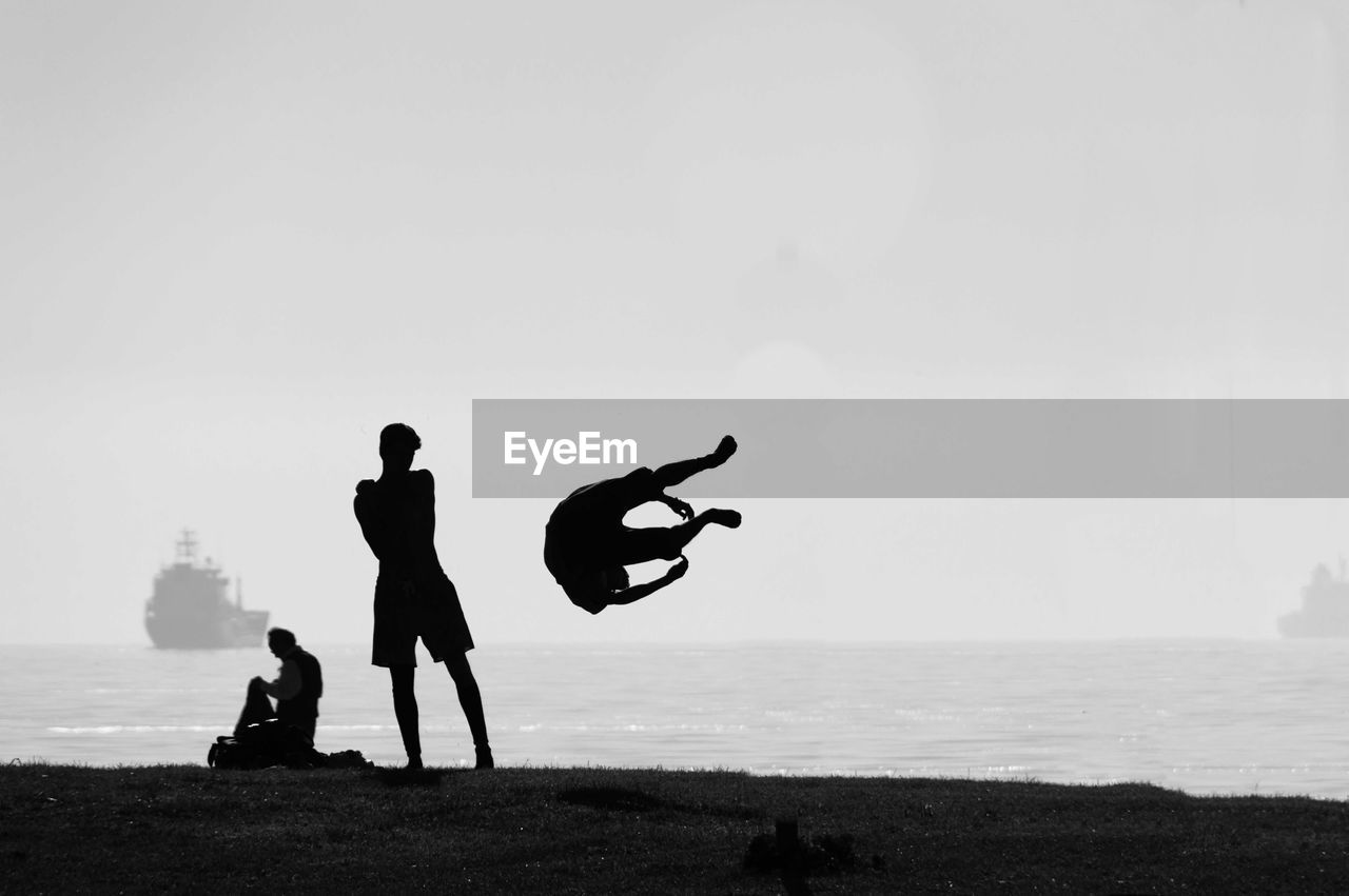 Silhouette man performing stunt at sea shore against sky