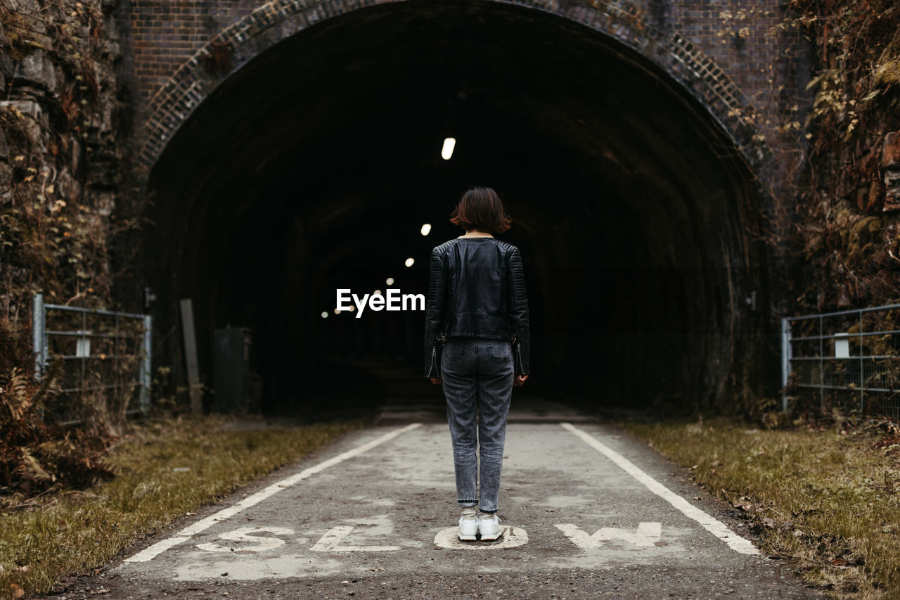 Back view of stylish woman in black leather jacket looking at dark tunnel with illumination inside on marked with inscription slow road in united kingdom