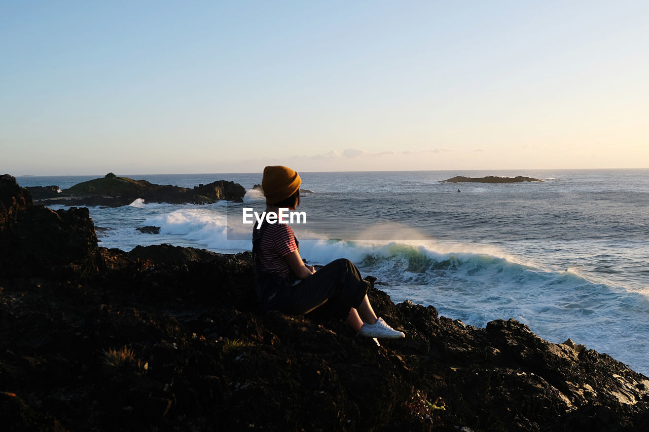 side view of woman sitting on rock at beach against sky