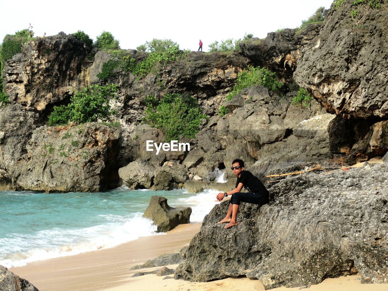 Side view of young man sitting on rock at beach against sky