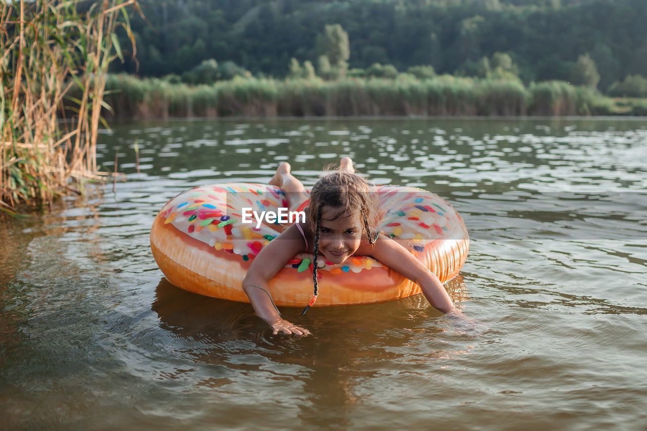 Girl has fun on big donut inflatable ring on lake on hot summer day, happy summertime, countryside