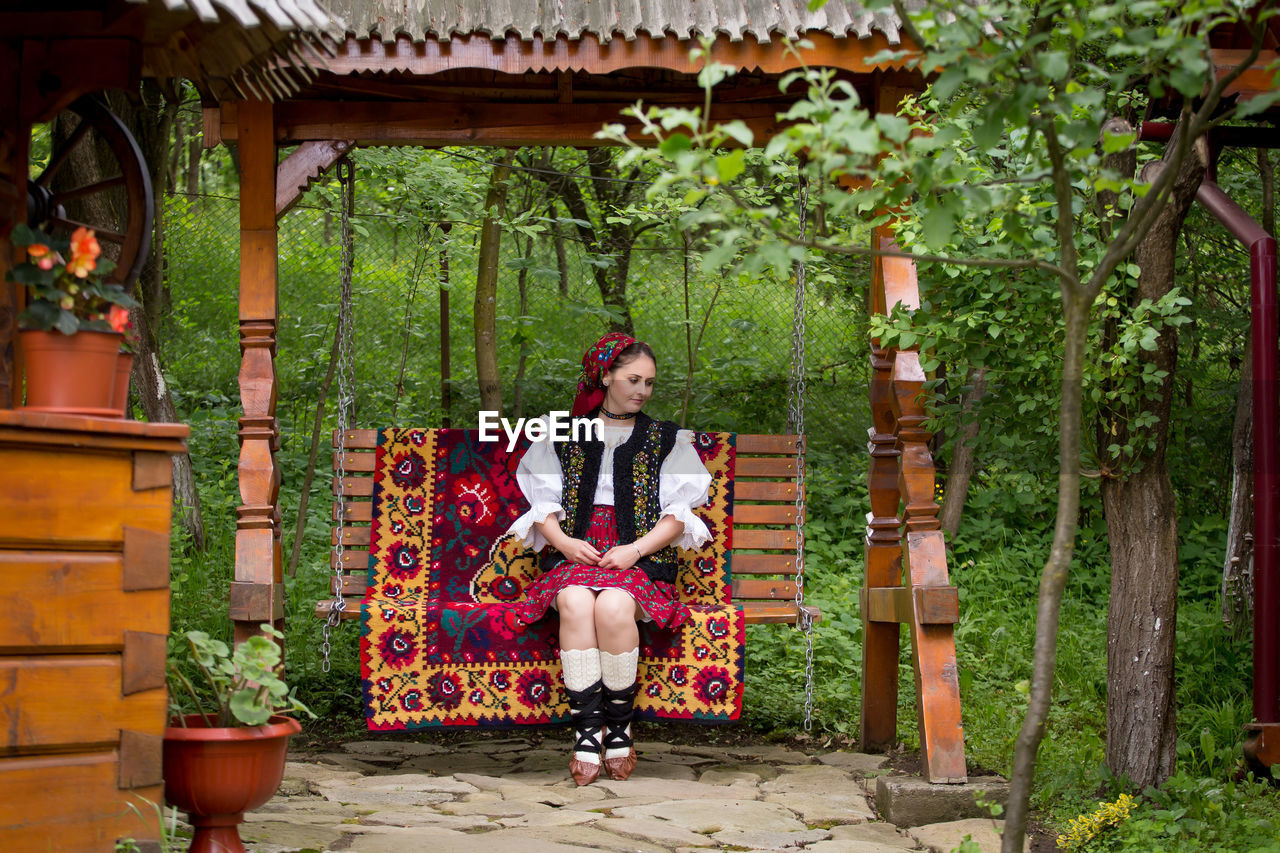 Full length of young woman in romanian clothing sitting on swing at yard