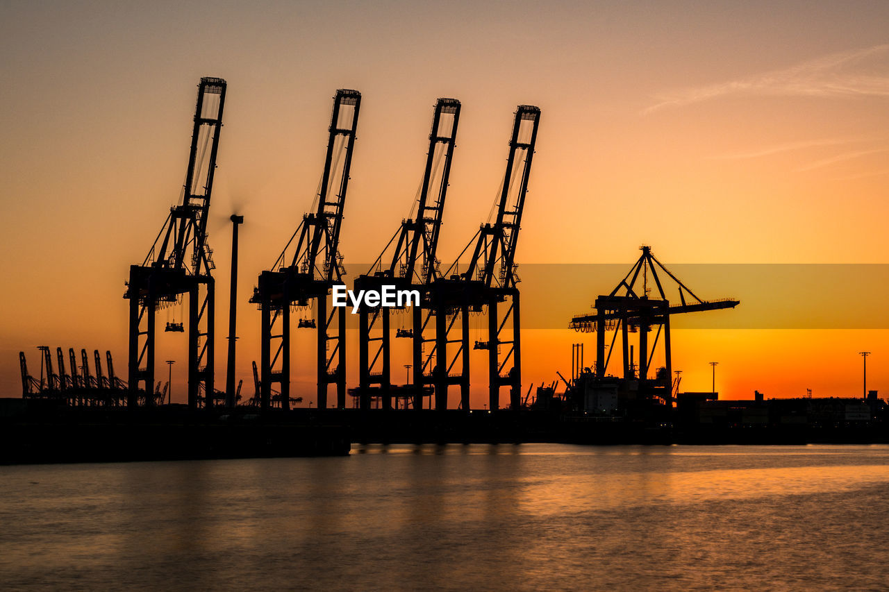 Silhouette cranes at commercial dock against sky during sunset