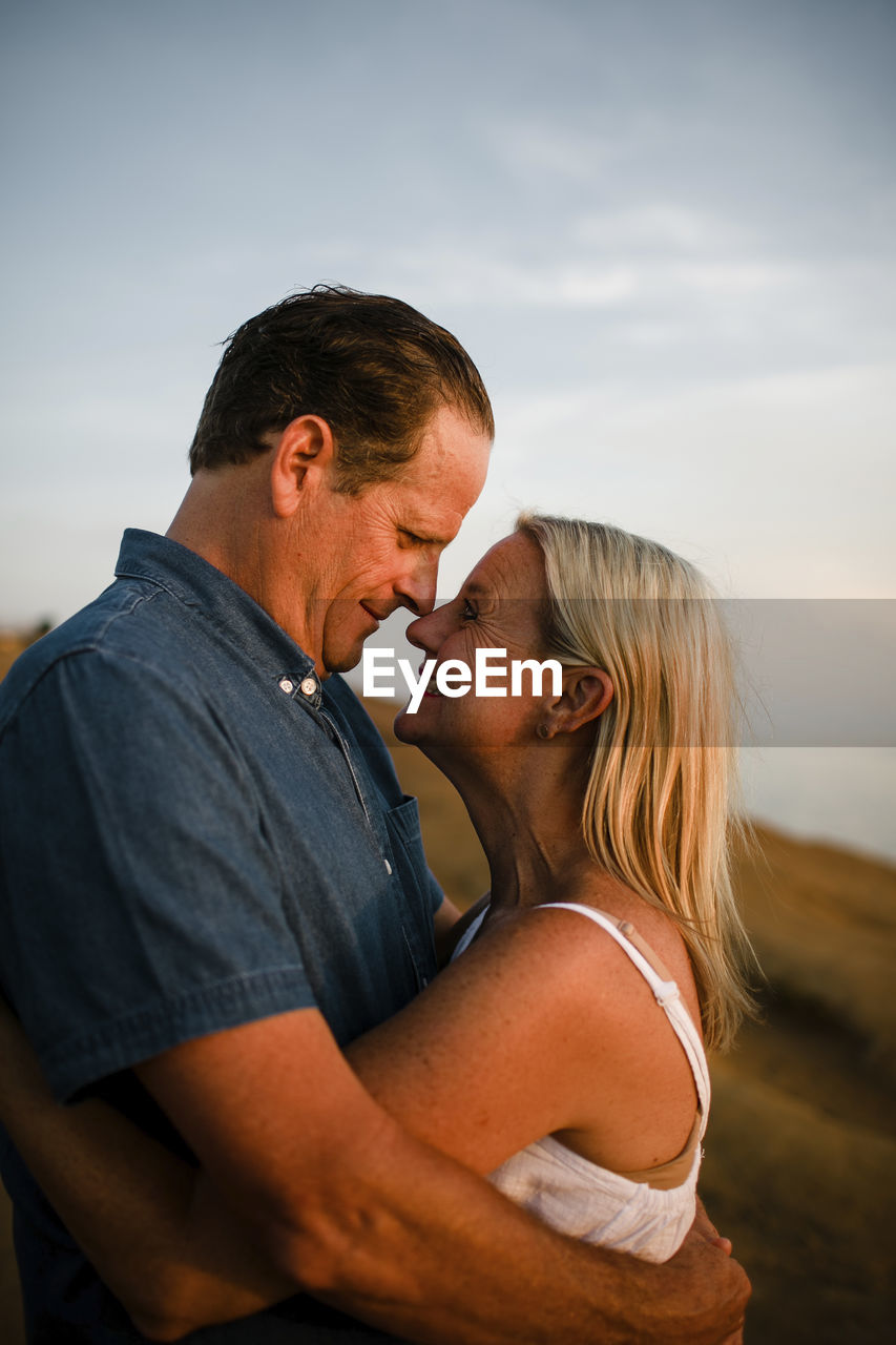 Side view of romantic couple looking at each other while standing on cliff against sky during sunset