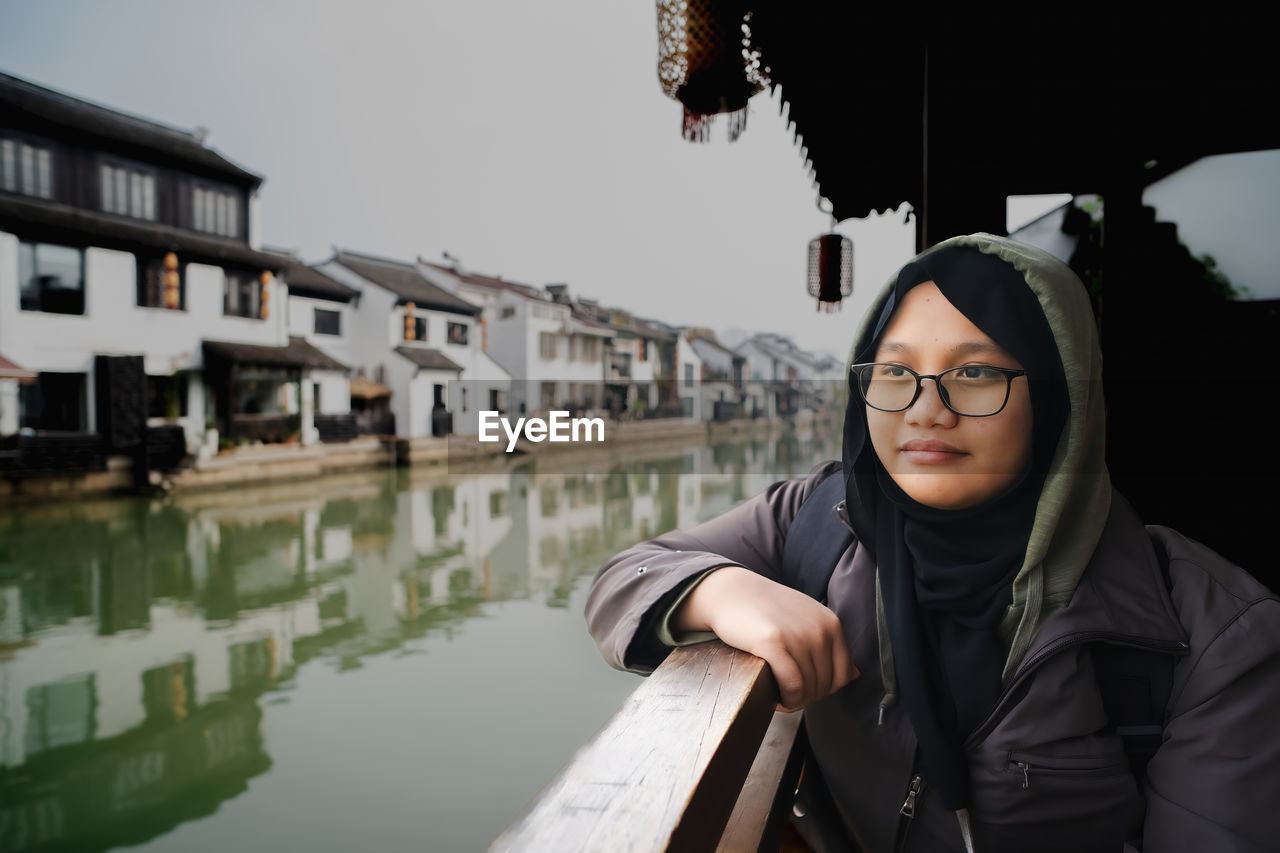 one person, architecture, water, glasses, adult, portrait, eyeglasses, young adult, built structure, reflection, building exterior, clothing, lifestyles, city, nature, women, canal, waist up, leisure activity, looking, outdoors, sky, looking at camera, standing, day, front view, casual clothing, building, headshot
