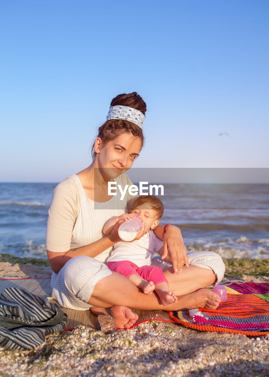 Portrait of smiling mother feeding milk to daughter while sitting at beach during sunset