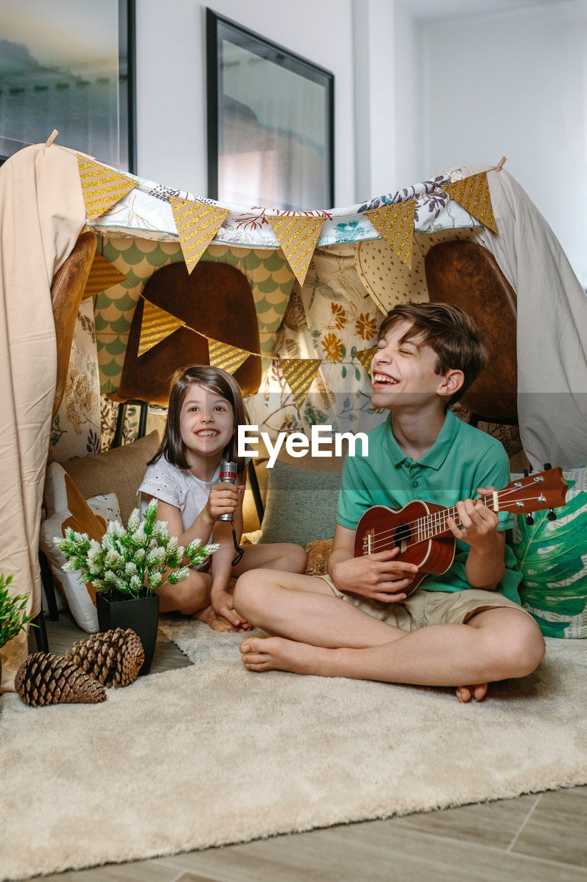 Happy children playing a ukulele and singing on handmade shelter tent in living room at home