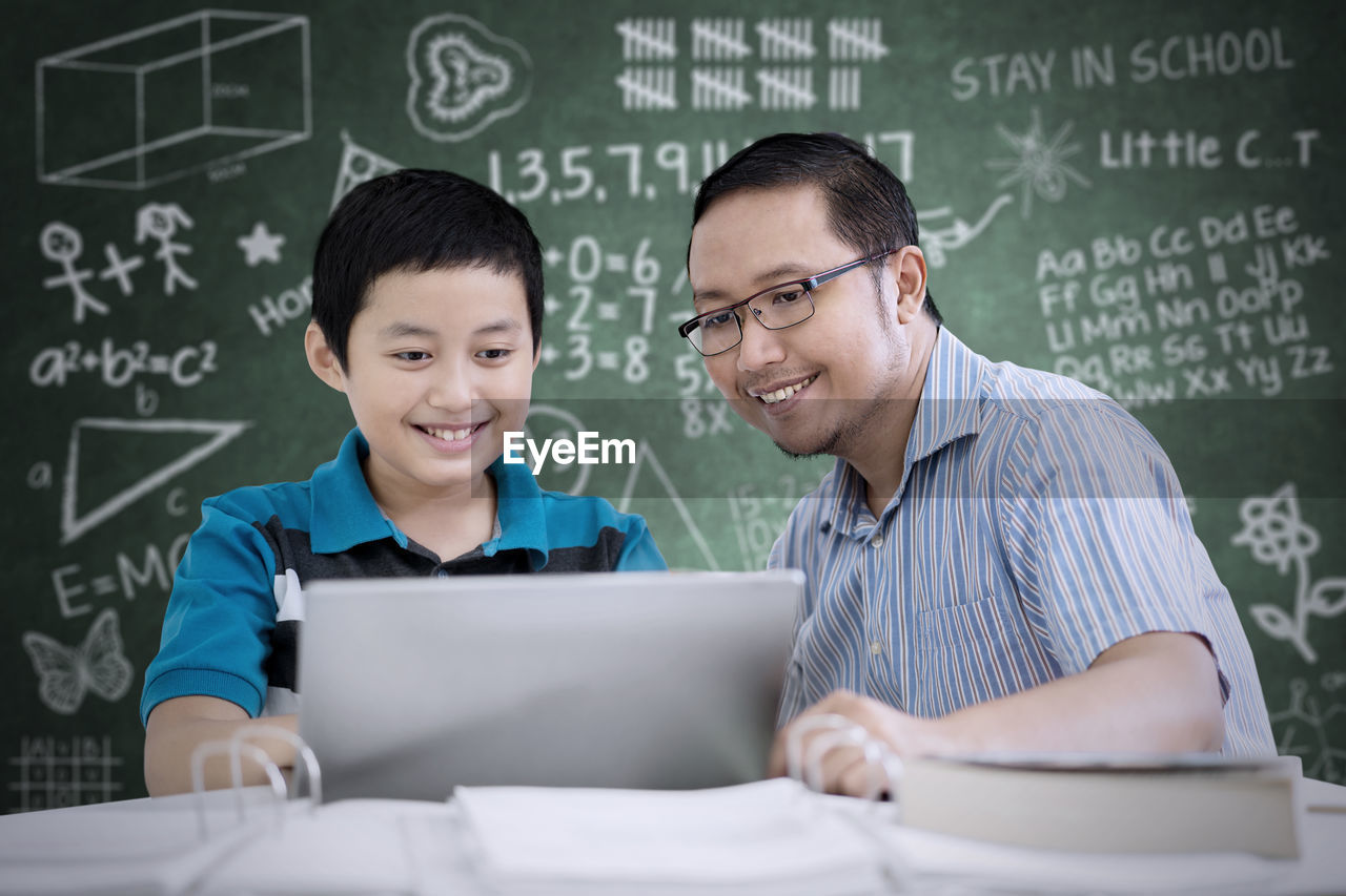 Boy with teacher at table against blackboard