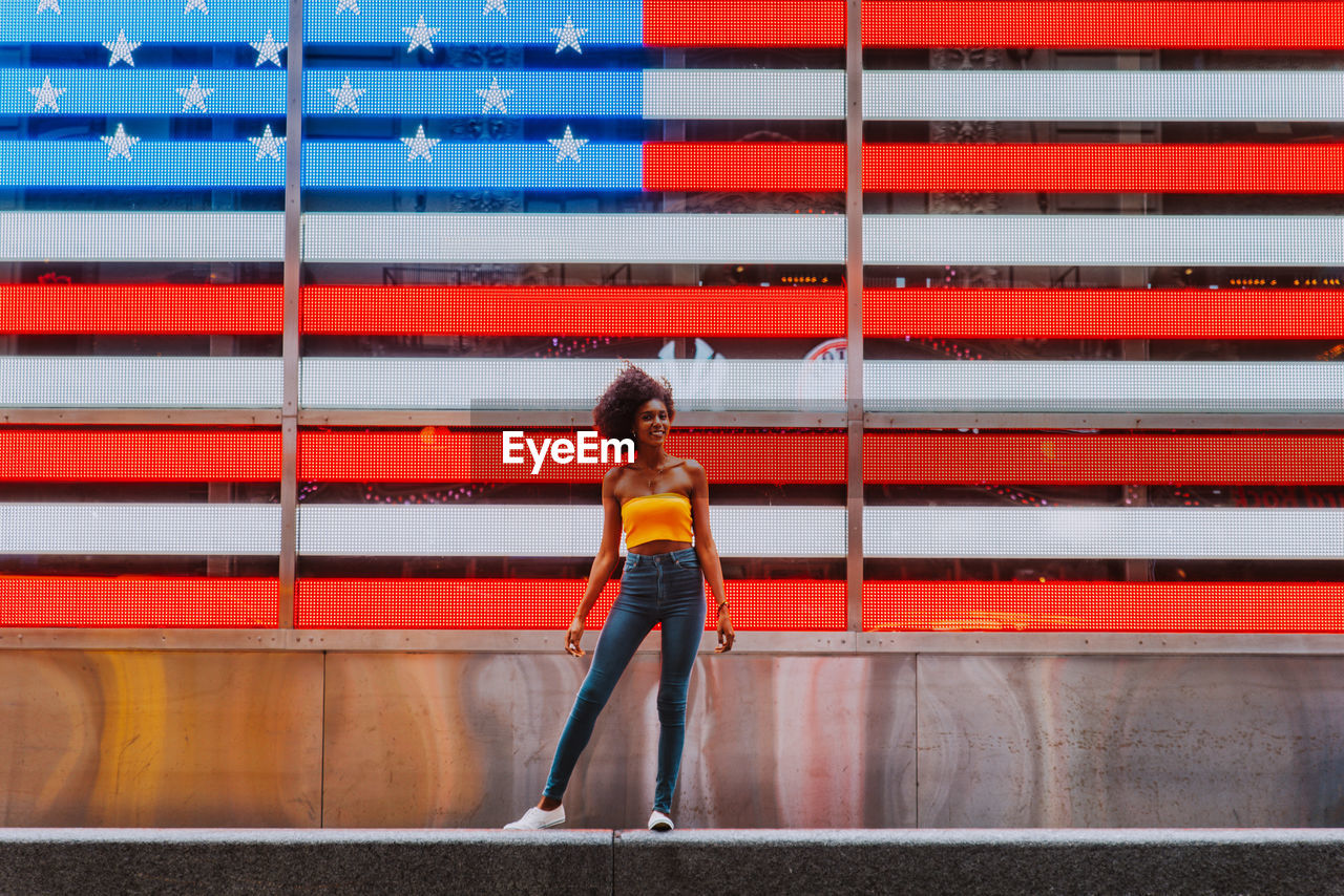 Full length portrait of happy young woman standing against neon american flag in city
