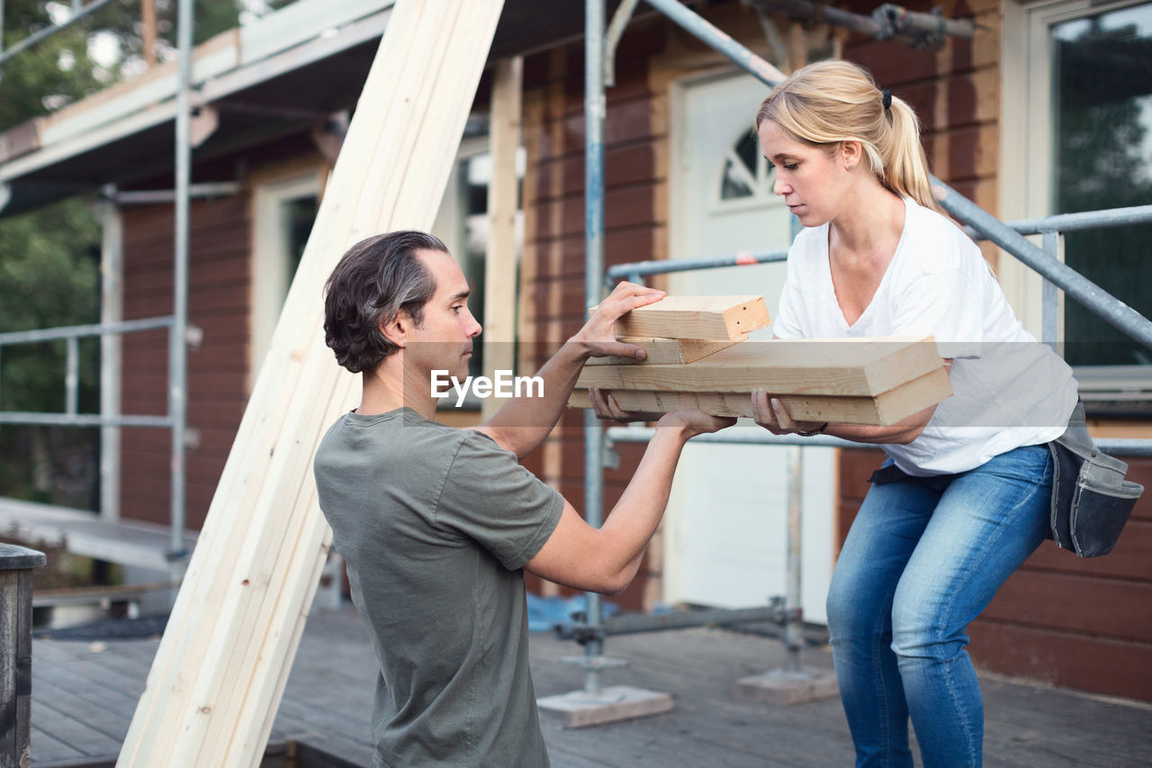 Side view of couple carrying stack of lumber outside house being renovated