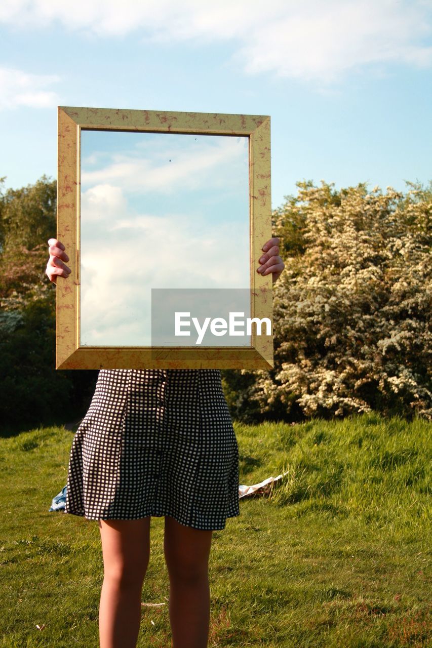 Midsection of woman holding picture frame with reflection of sky on land