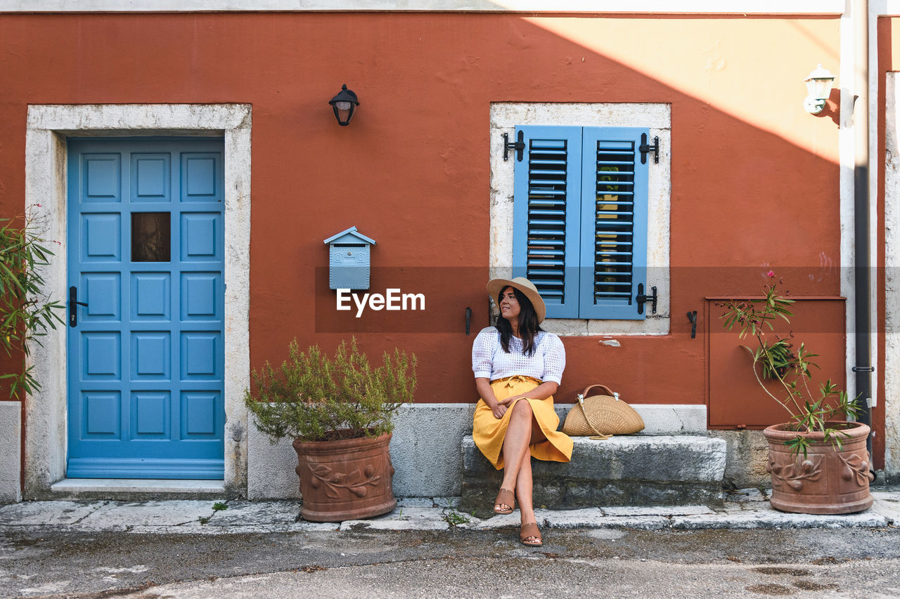 Young woman in summer outfit sitting on bench in front of colorful house.