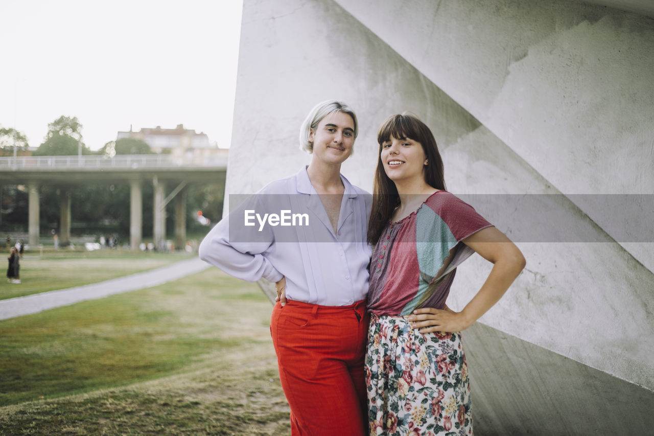 Portrait of smiling non-binary person and woman standing with hands on hips at park