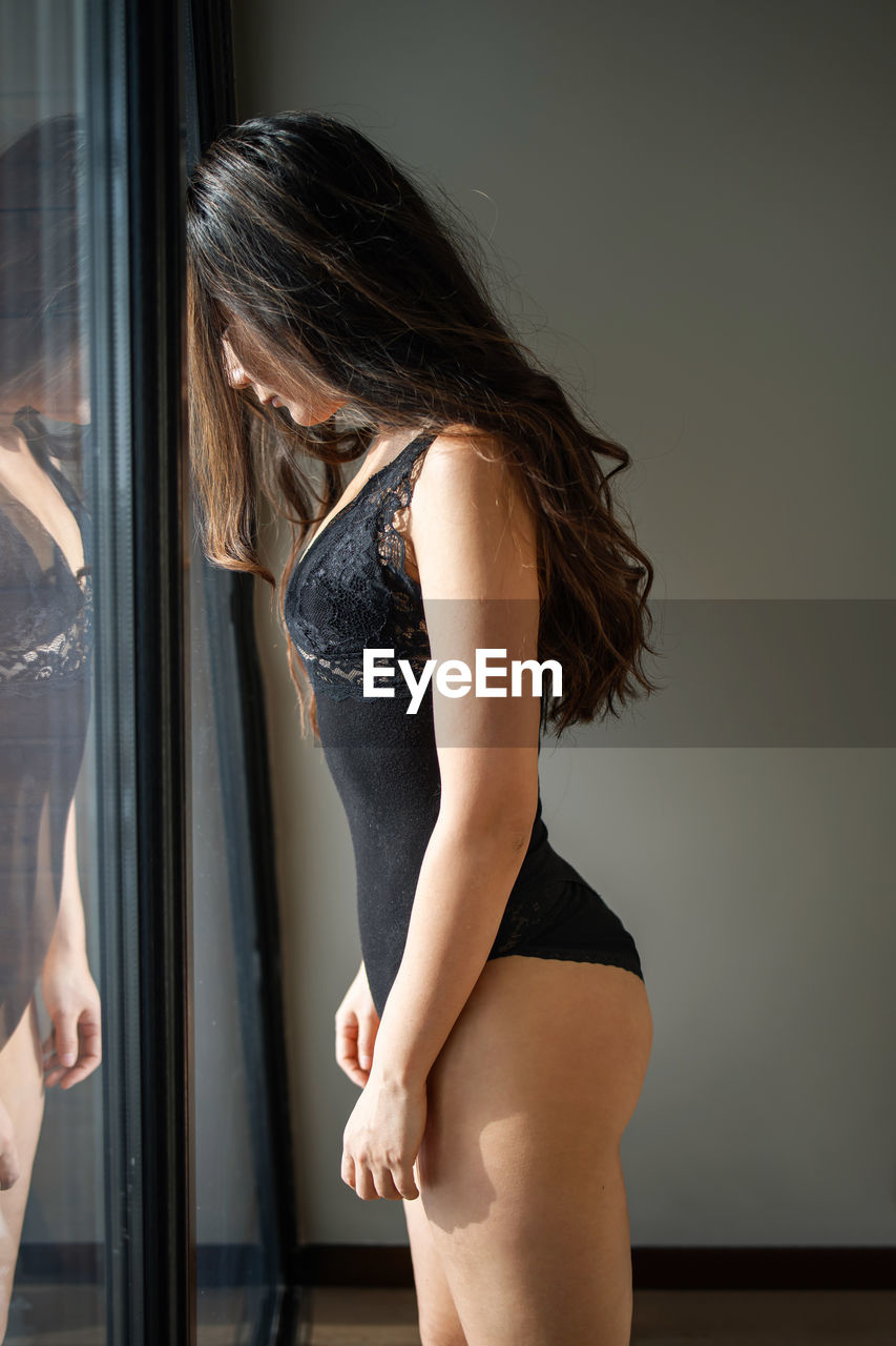 Sad depressed asian woman wearing attractive black lingerie standing near window looking down.