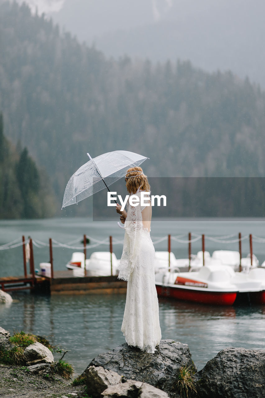 Beautiful young woman bride in a boho dress and with an umbrella stands in the rain in nature