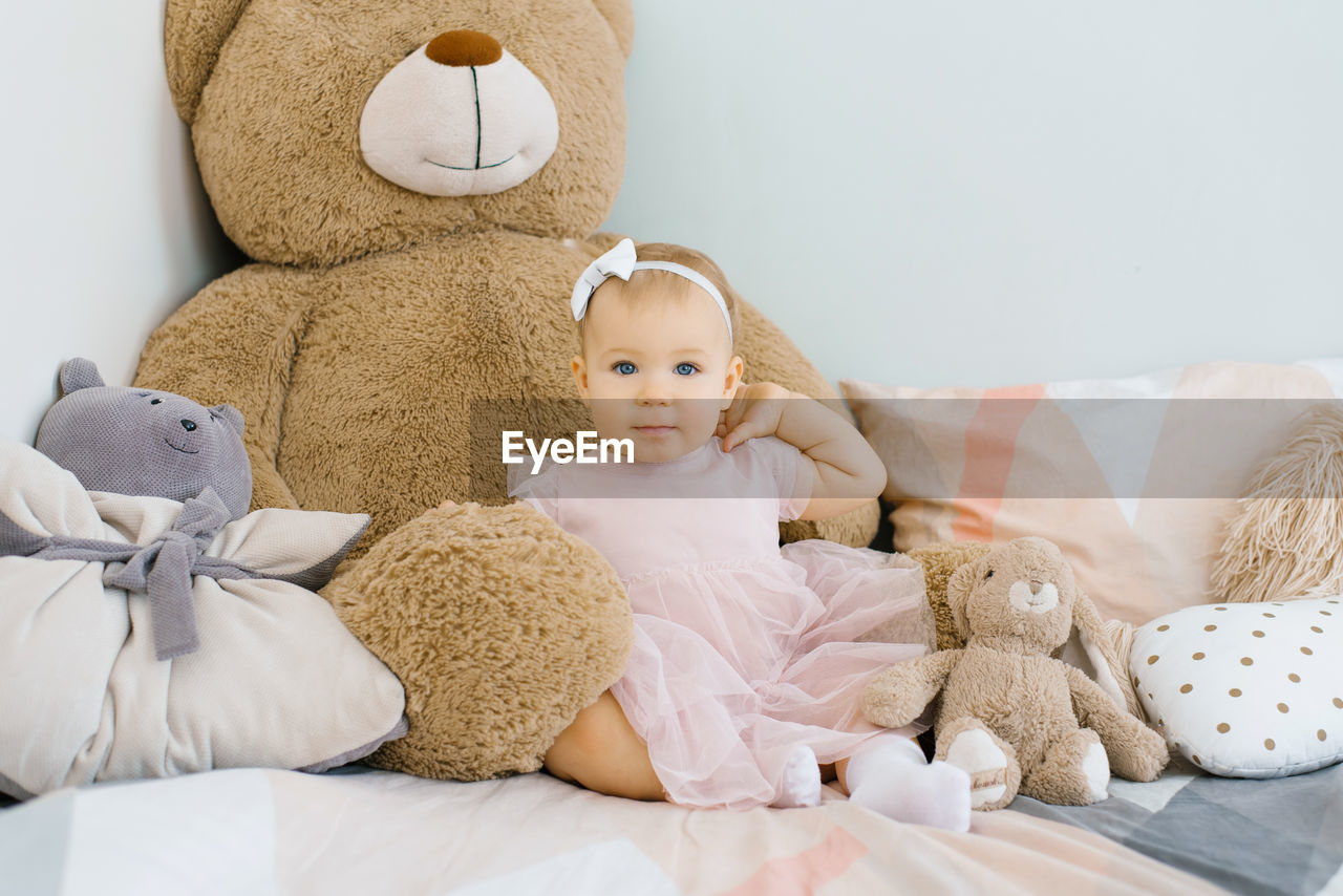 A cute one year old baby sits surrounded by soft animals toys and pillows