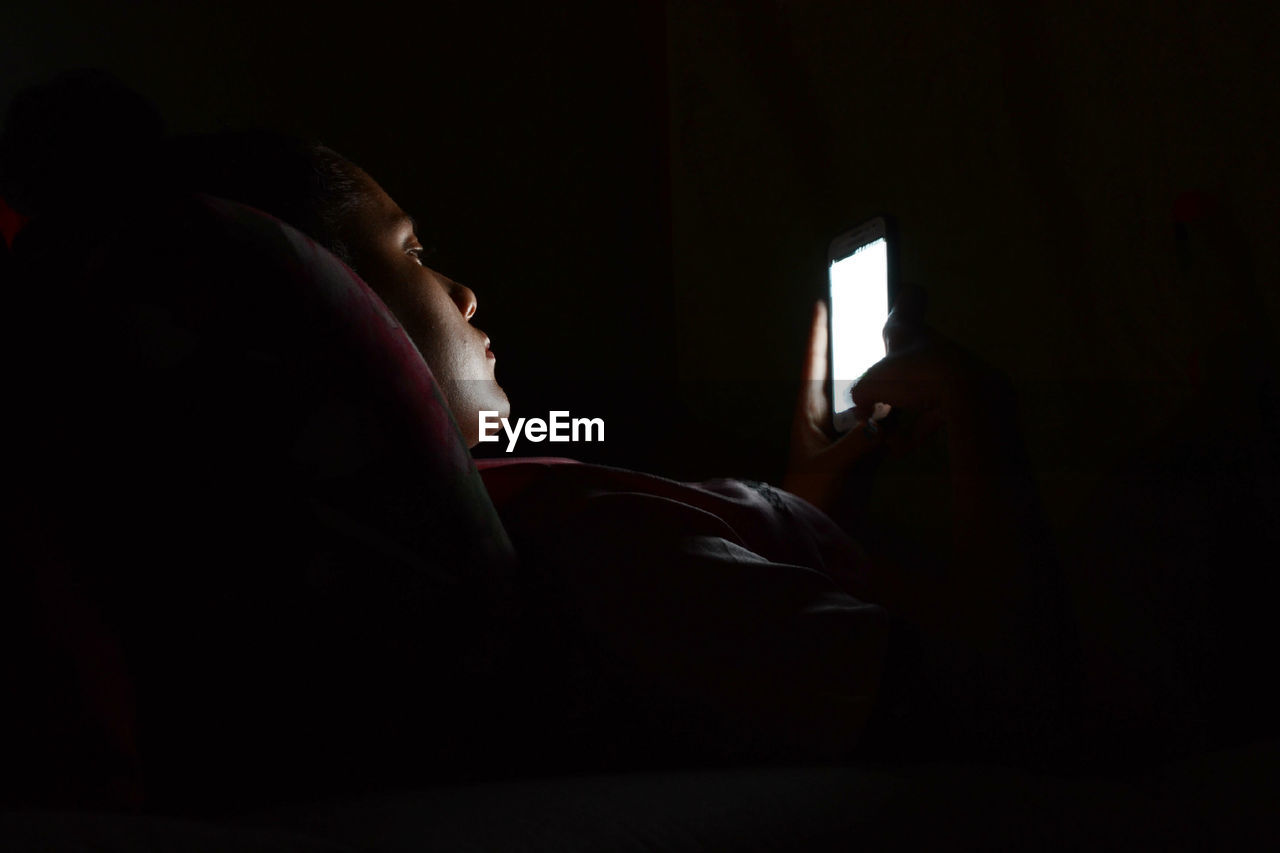 Side view of woman using phone while lying in darkroom