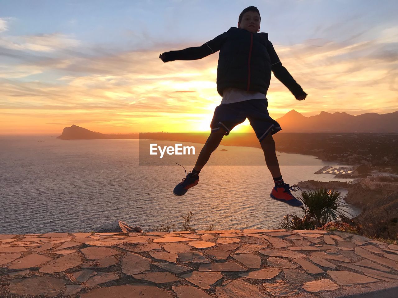 FULL LENGTH OF MAN JUMPING ON SEA SHORE AGAINST SKY DURING SUNSET