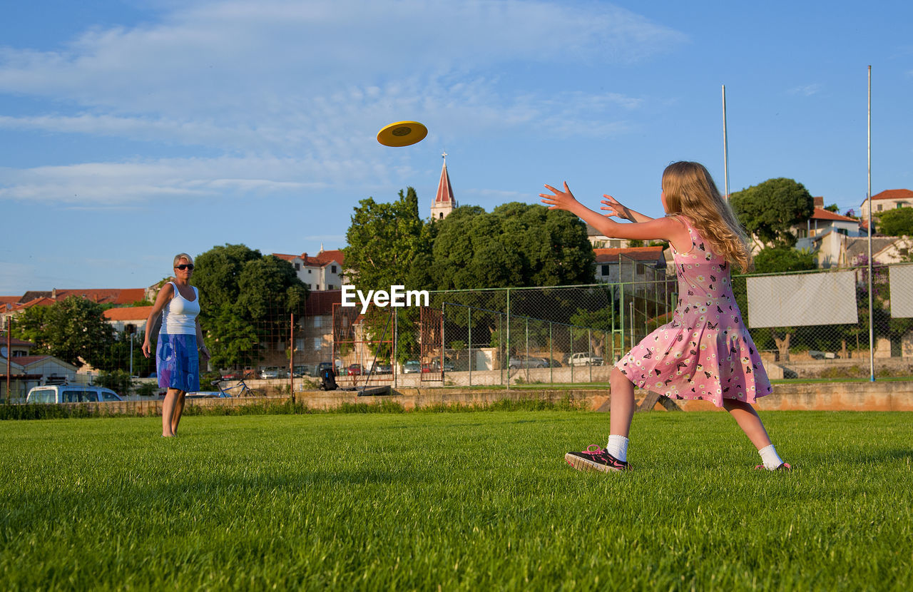 Woman and girl playing with plastic disc on grass against sky