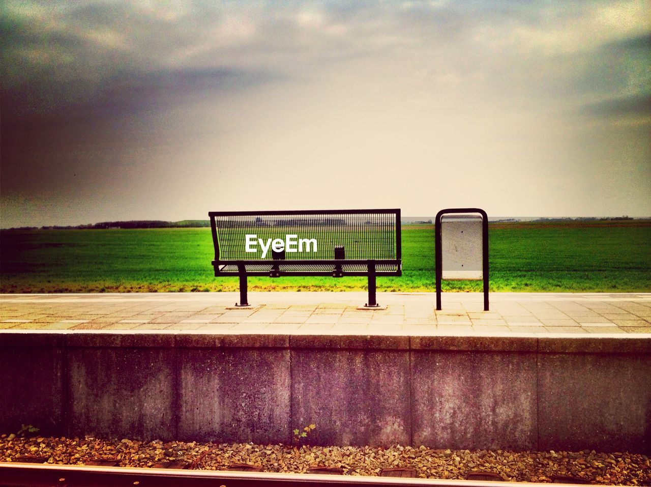 Empty bench by garbage can on railroad station platform against cloudy sky