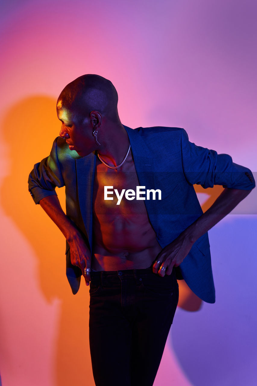 Confident bald african american slim male model with necklace in trendy outfit standing at wall with glowing lights in modern studio