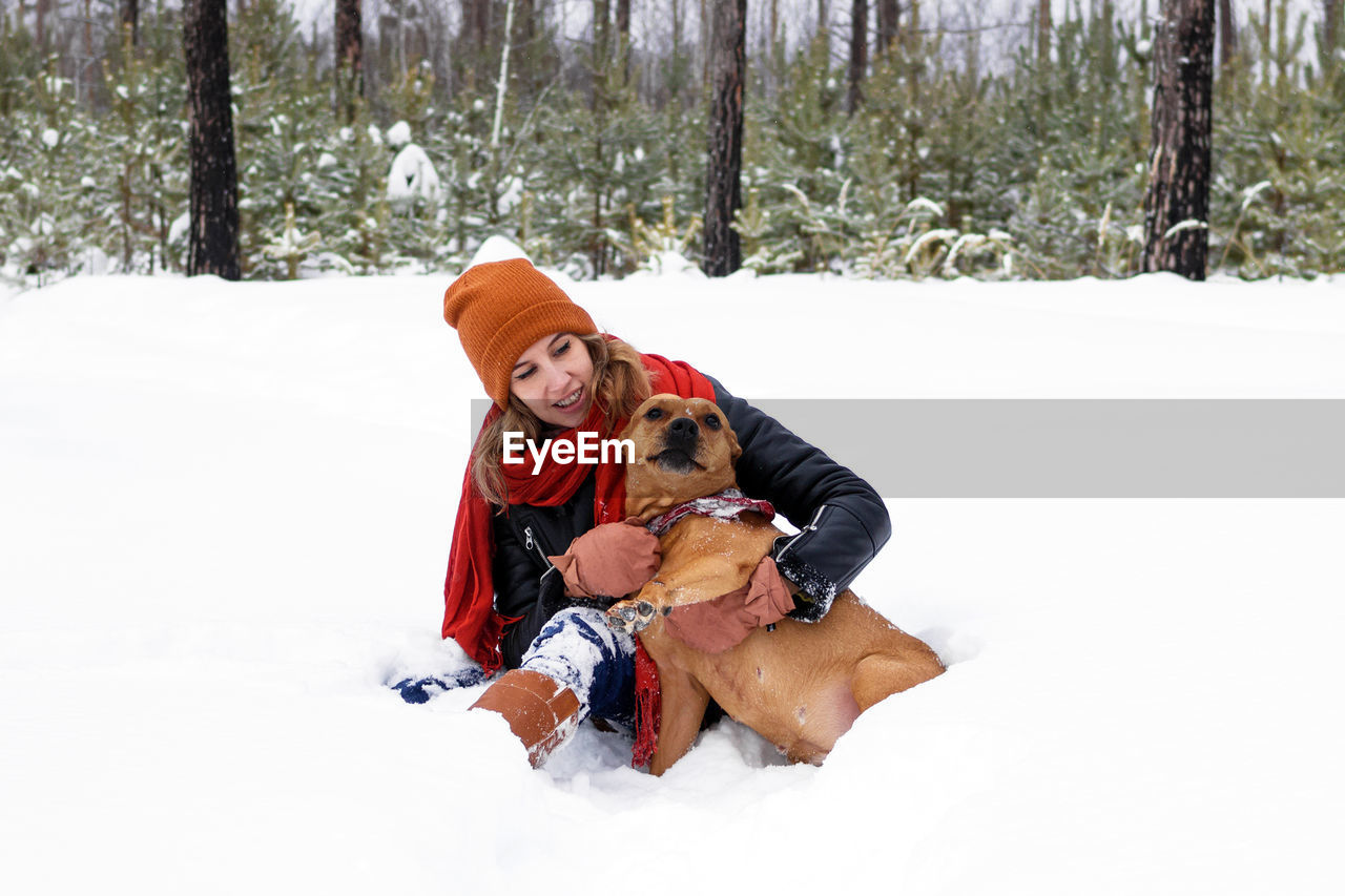 Beautiful young woman is sitting on a snow and playing with her dog in winter coniferous forest.