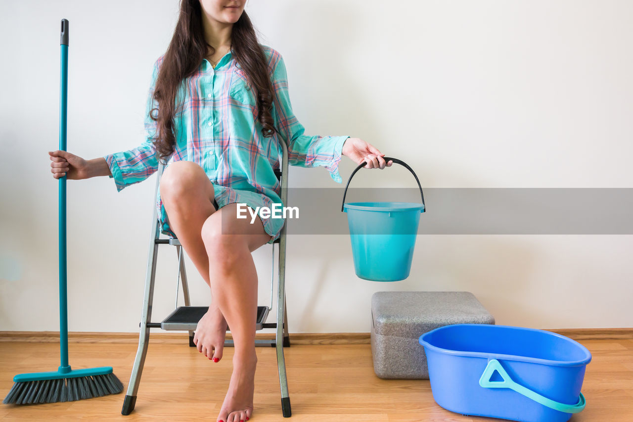 Low section of woman holding bucket and mop while sitting on ladder against wall at home