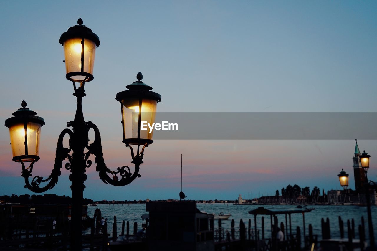 Illuminated street light by sea against sky during sunset