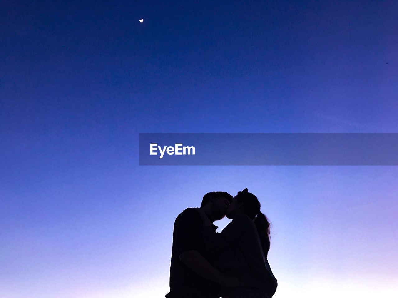 Low angle view of couple kissing while standing against blue sky during sunset