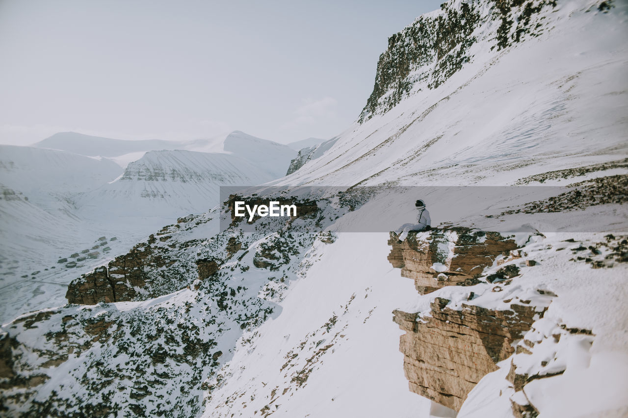 Unrecognizable cosmonaut wearing white spacesuit sitting on edge of rocky mountain in winter and admiring amazing landscape in svalbard