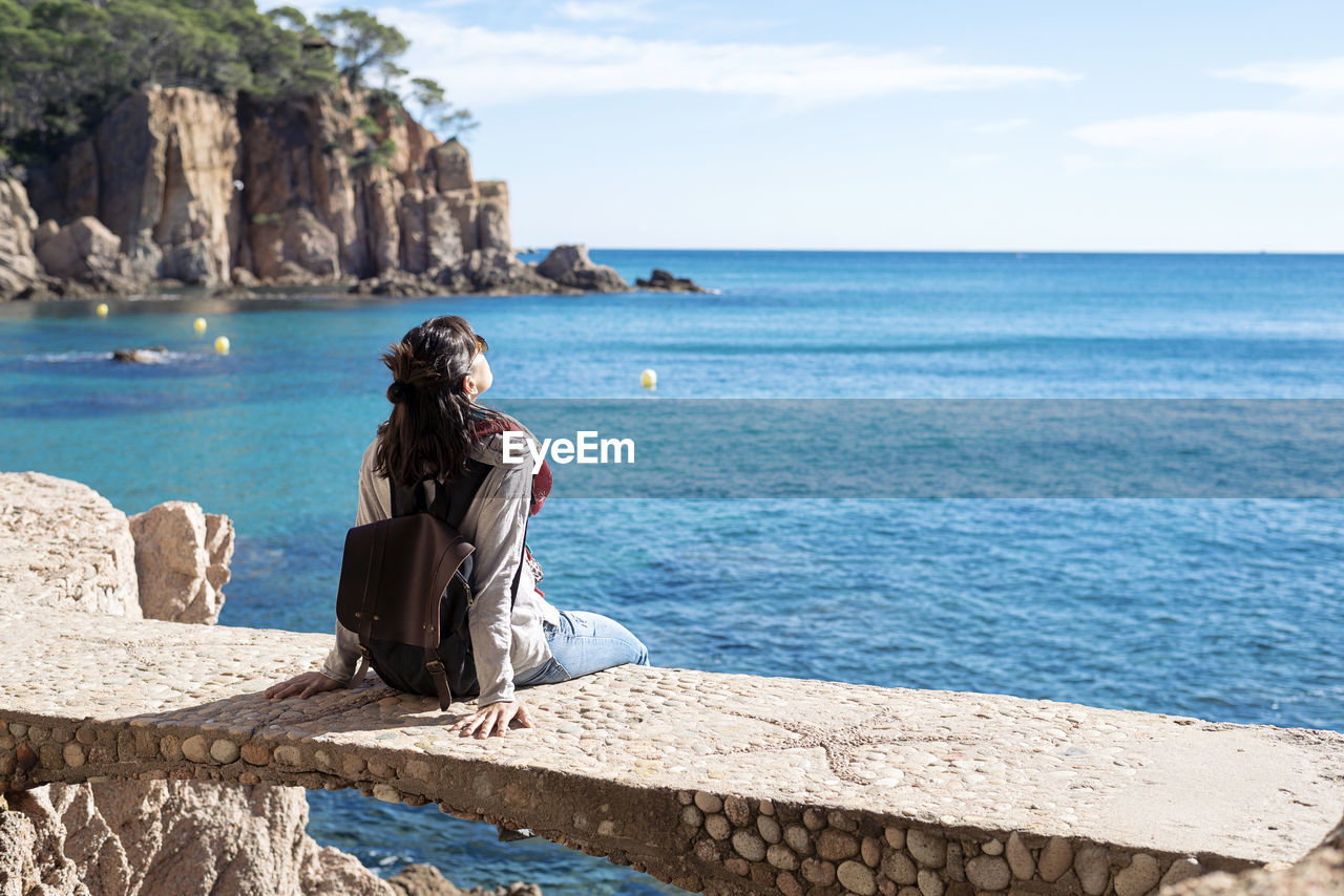 Rear view of a woman relaxing in front of blue sea, sitting on bridge