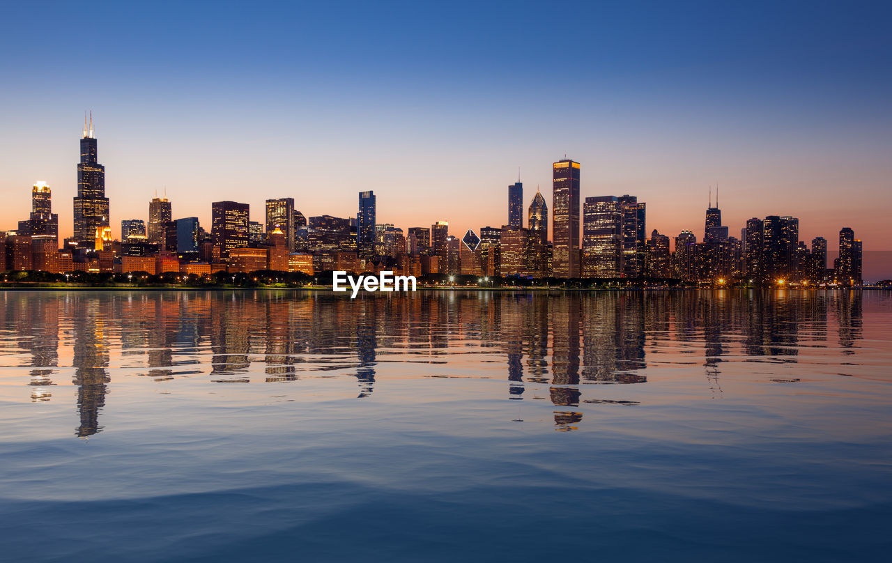 Illuminated buildings by lake in city against sky at dusk