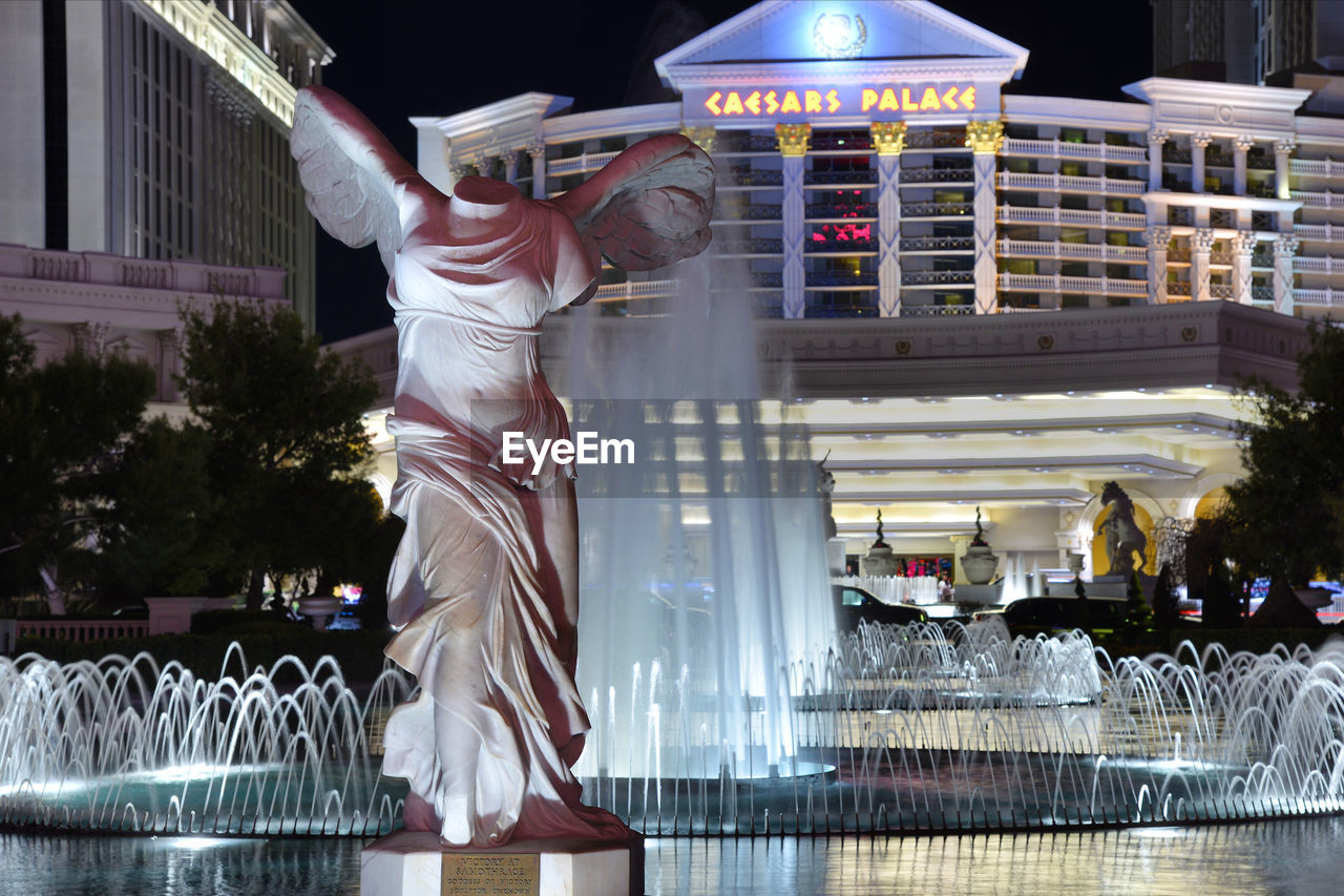 LOW ANGLE VIEW OF WOMAN WITH ILLUMINATED SCULPTURE AGAINST CITY AT NIGHT