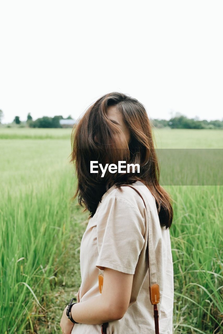 Woman covering face with brown hair standing amidst plants on field