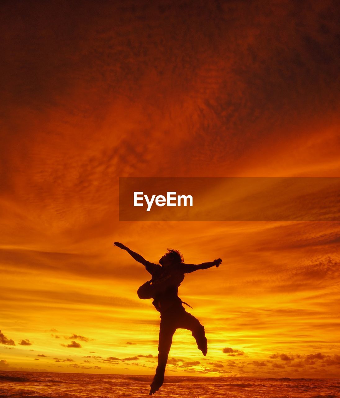 Silhouette man with arms outstretched jumping against orange sky