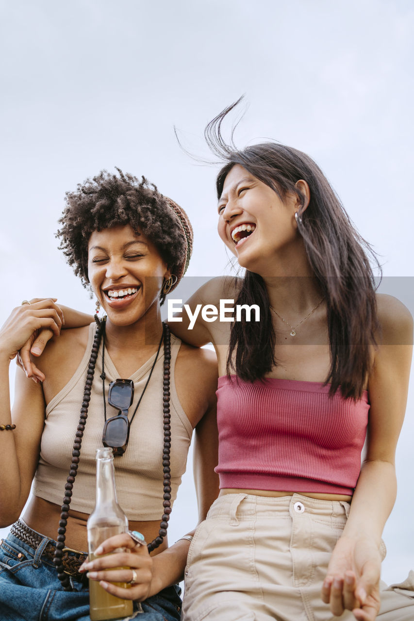 Cheerful young women laughing while sitting against sky