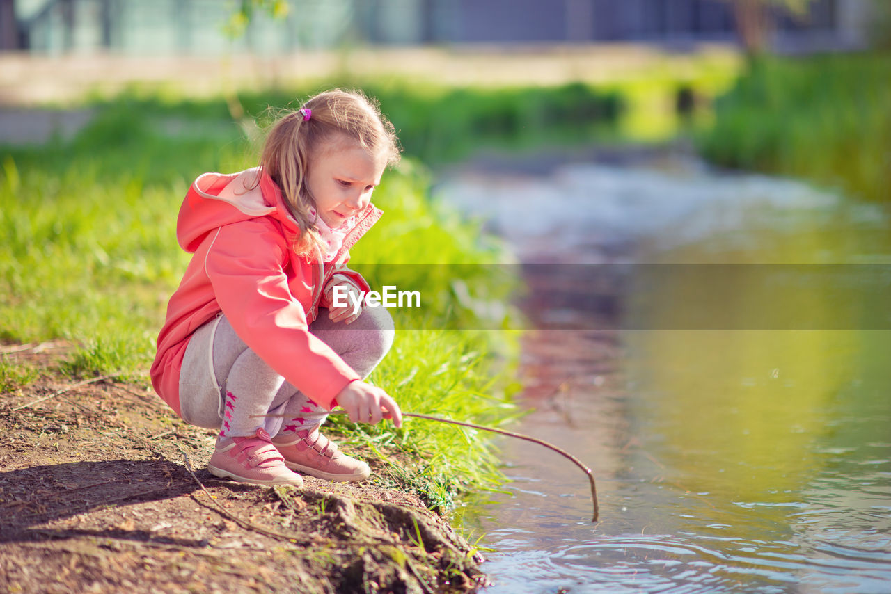 Girl holding stick while crouching by canal