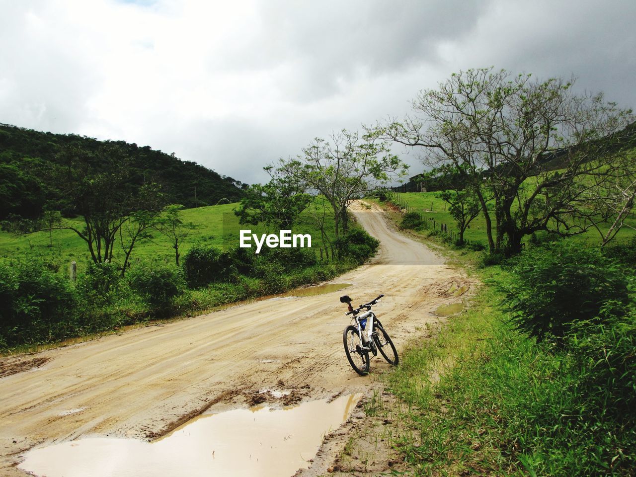 Bicycle parked by puddle in dirt road against cloudy sky