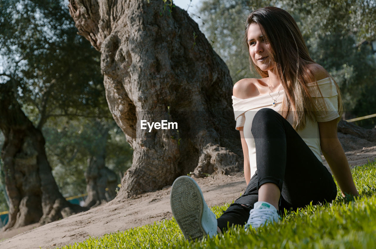 Tilt image of smiling young woman sitting against trees in park