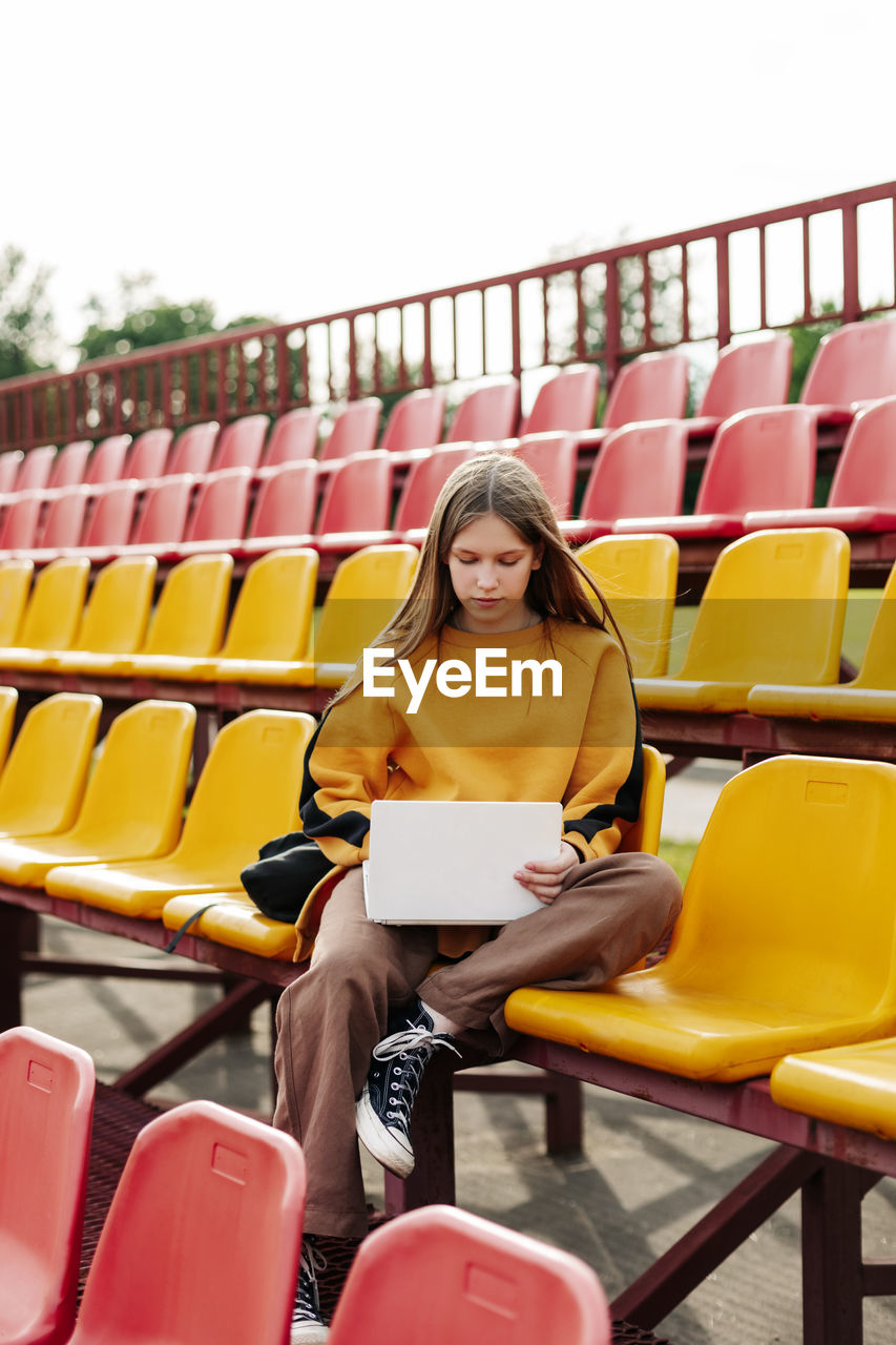 A young girl is doing her homework using a laptop in the stands of the stadium.