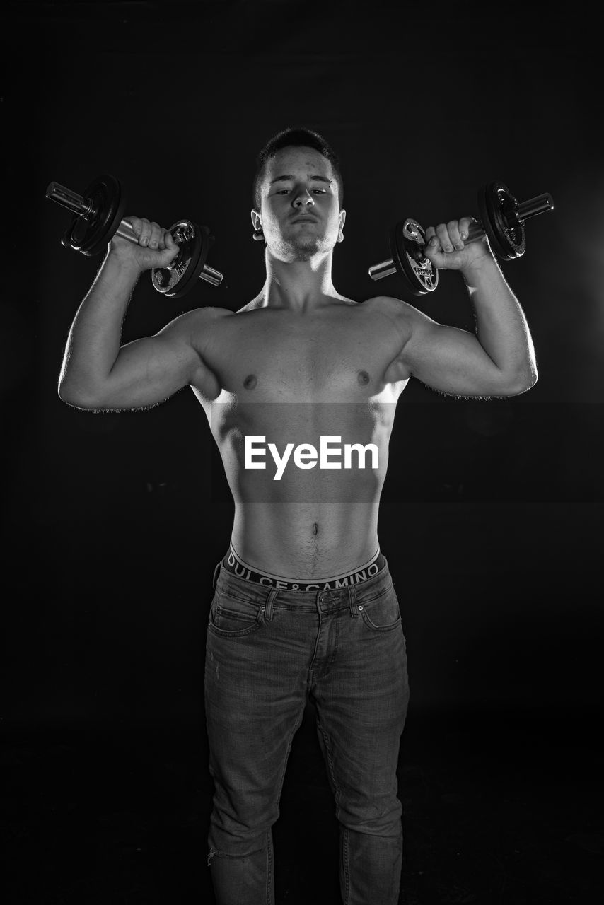 Portrait of shirtless man lifting dumbbells against black background