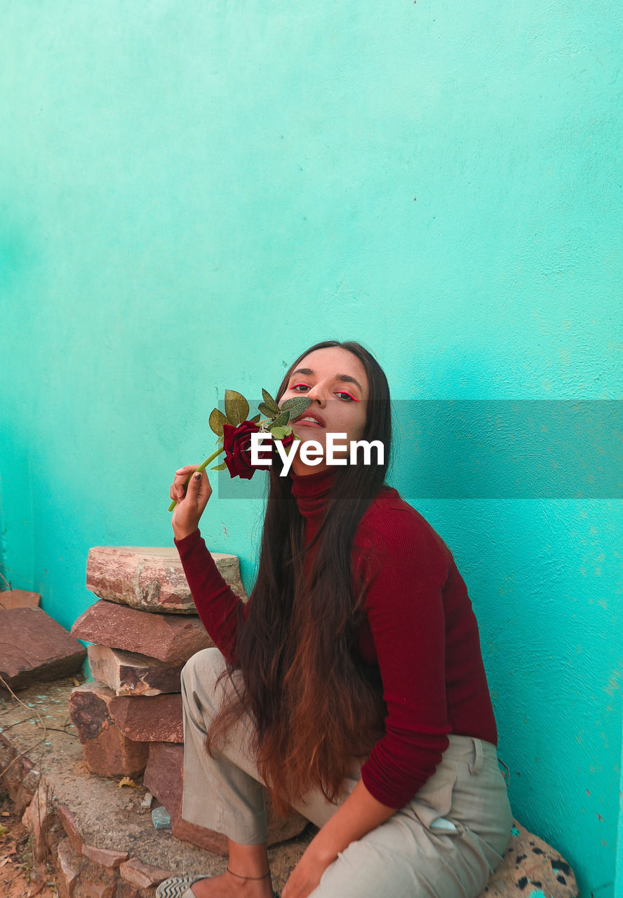 Portrait of young woman holding rose while crouching against wall