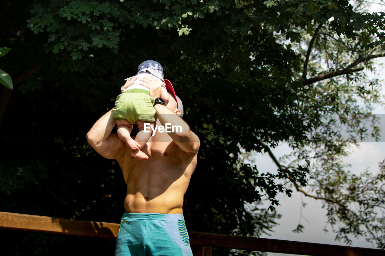 Shirtless father playing with son while standing against branches