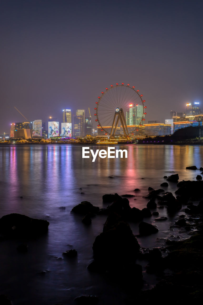 ILLUMINATED FERRIS WHEEL BY SEA AGAINST SKY IN CITY