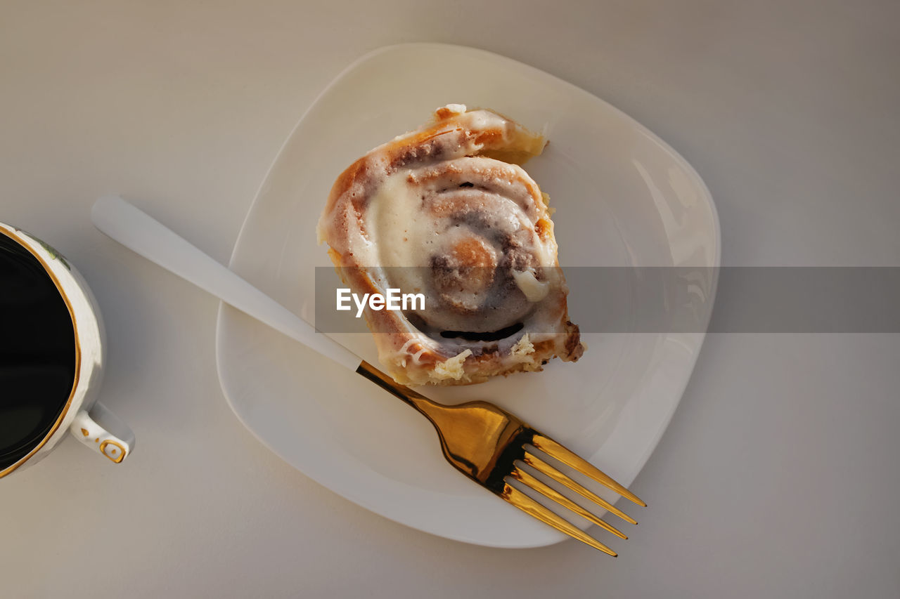 Cinnamon bun on a white plate and elegant fork on the white table, top view. 