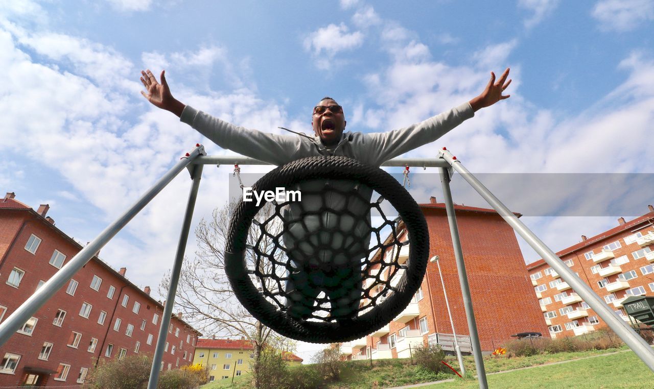 Low angle view of man with arms outstretched screaming on swing against sky